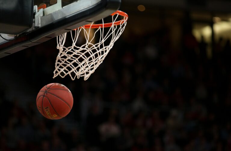 A low-angle, close-up shot of an orange basketball just about to go through a white net and rim in a dimly lit indoor arena. The background is dark and blurry, suggesting a crowd of spectators in the stands.