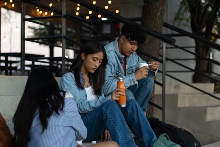Three young adults sitting on concrete outdoor stairs at dusk, dressed in casual denim. A woman in the center holds an orange water bottle, while a man next to her eats from a white container. String lights and a black metal railing are visible in the background.