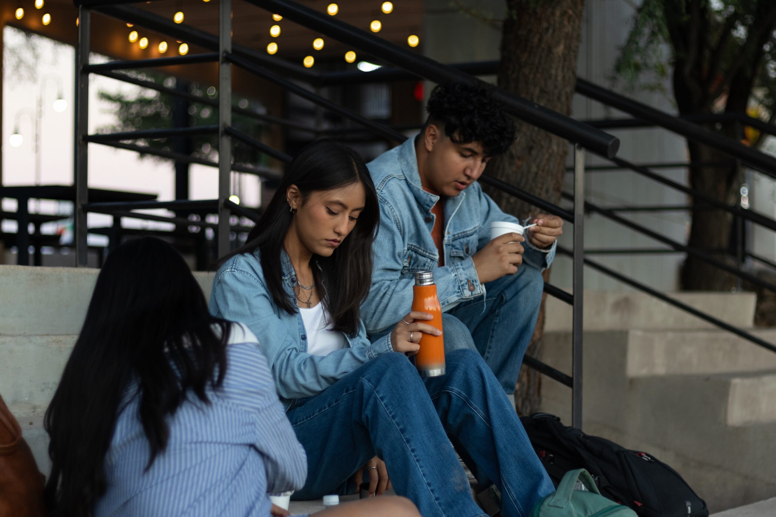 Three young adults sitting on concrete outdoor stairs at dusk, dressed in casual denim. A woman in the center holds an orange water bottle, while a man next to her eats from a white container. String lights and a black metal railing are visible in the background.