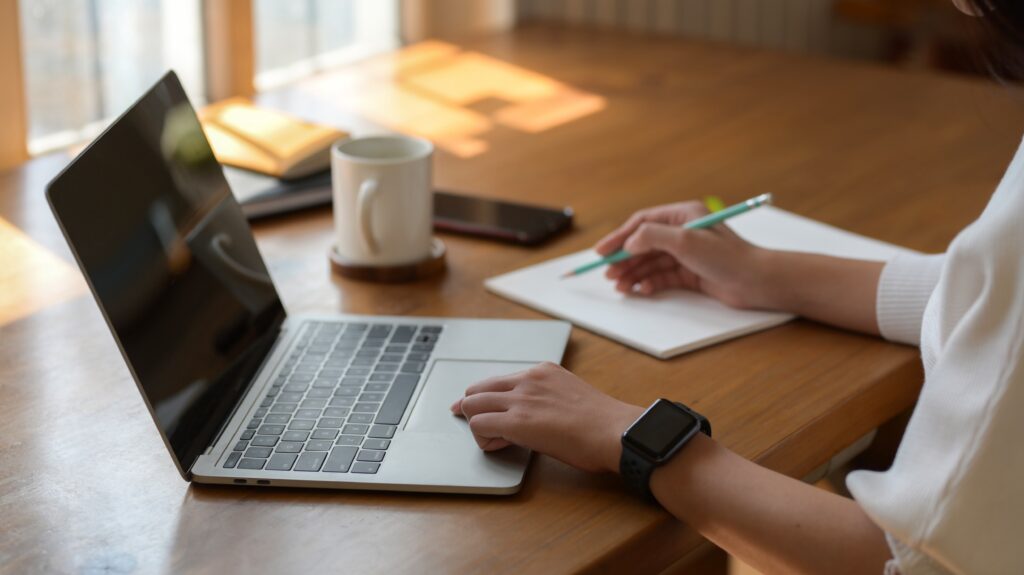 young college girl working on laptop while writing on notebook in simple workspace