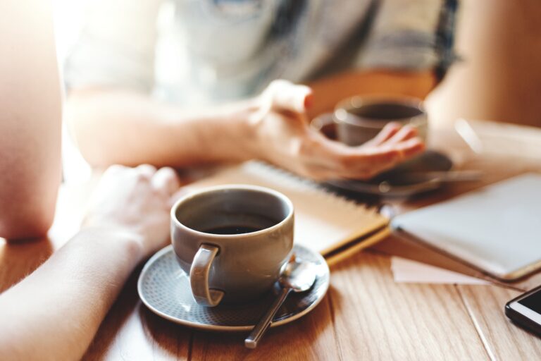 Friends talking at cafe table during coffee break. Unrecognizable male and female colleagues discussing business issues, focus on coffee cup with saucer and teaspoon